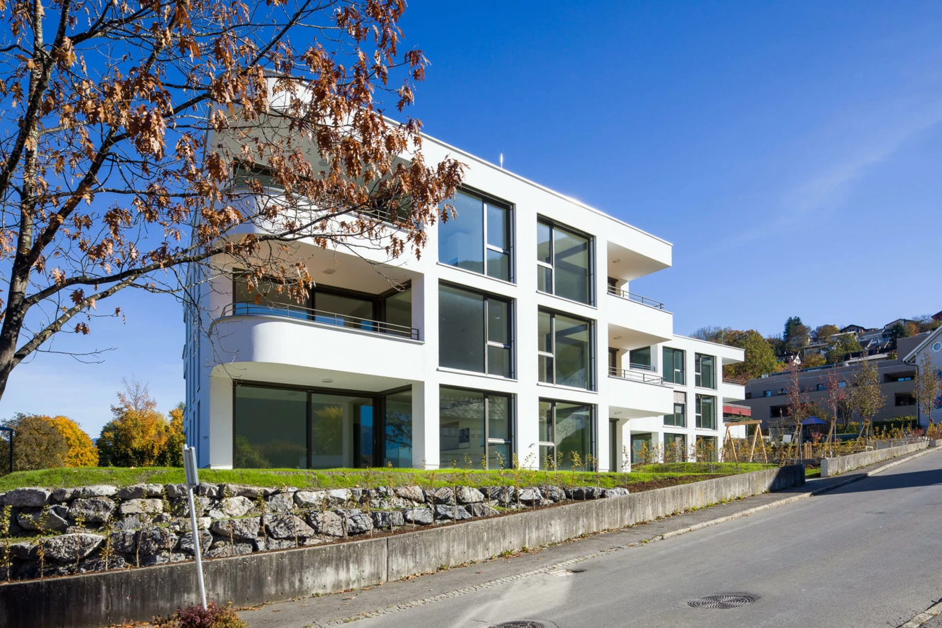 Moderne Wohnanlage Wolf-Huber-Straße in Feldkirch mit weißer Fassade, großen Fenstern und Balkonen an einem sonnigen Herbsttag.