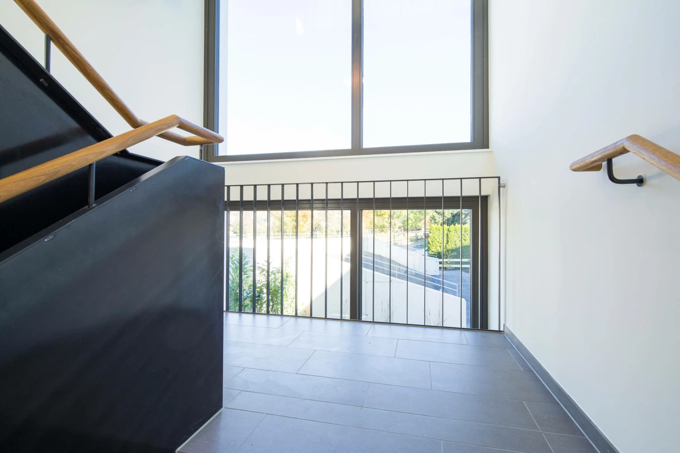 Moderner Treppenbereich mit dunkler Treppe, Holzhandläufen und großer Fensterfront in der Wohnanlage Wolf-Huber-Straße, Feldkirch.