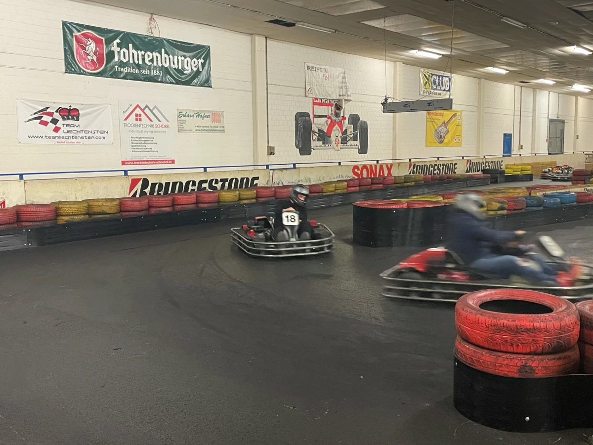 Zwei Personen fahren Go-Karts auf einer Rennstrecke mit Reifenstapeln und Werbebannern an den Wänden (Lehrlingsabend).