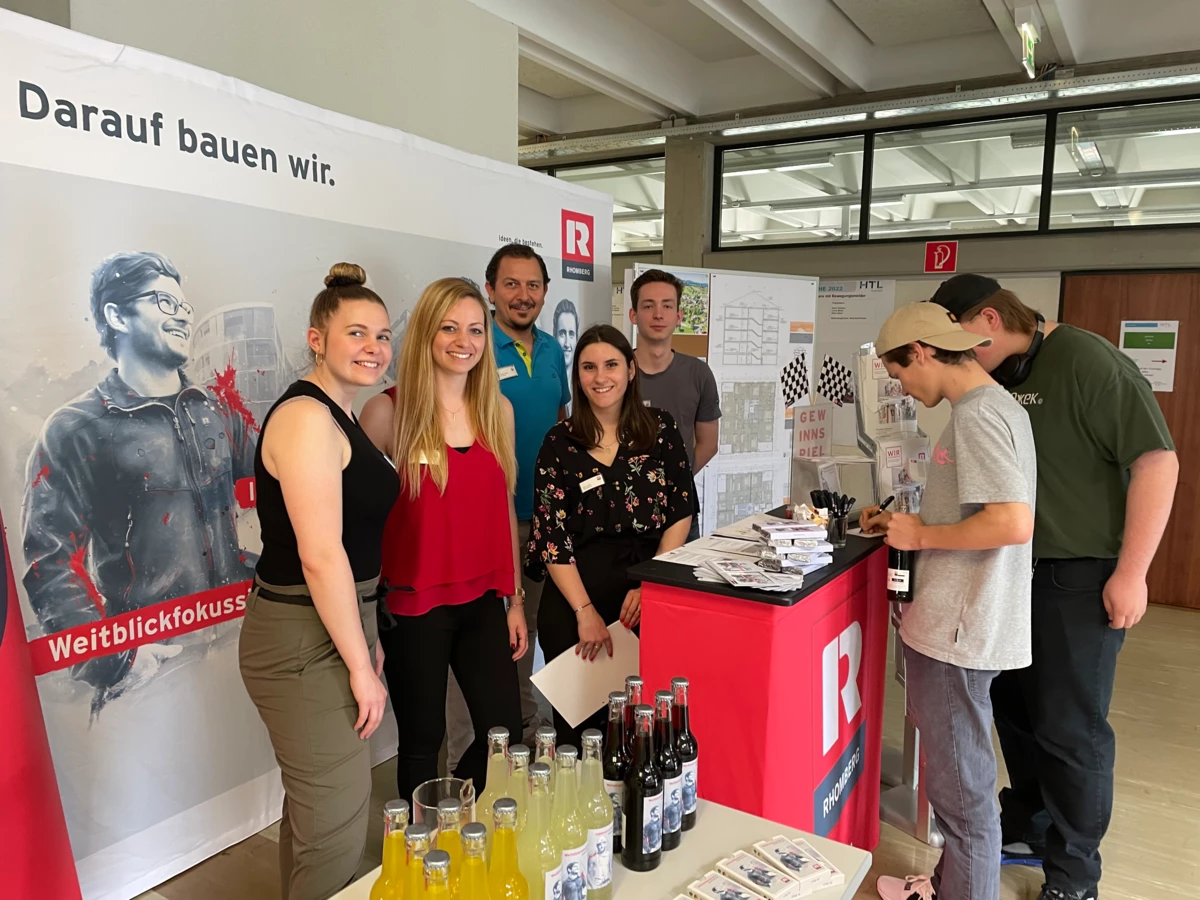 Mitarbeiter von Rhomberg Bau an einem Messestand, der "Darauf bauen wir" und "Weitblickfokus" bewirbt mit Getränken und Broschüren (Bauforum).