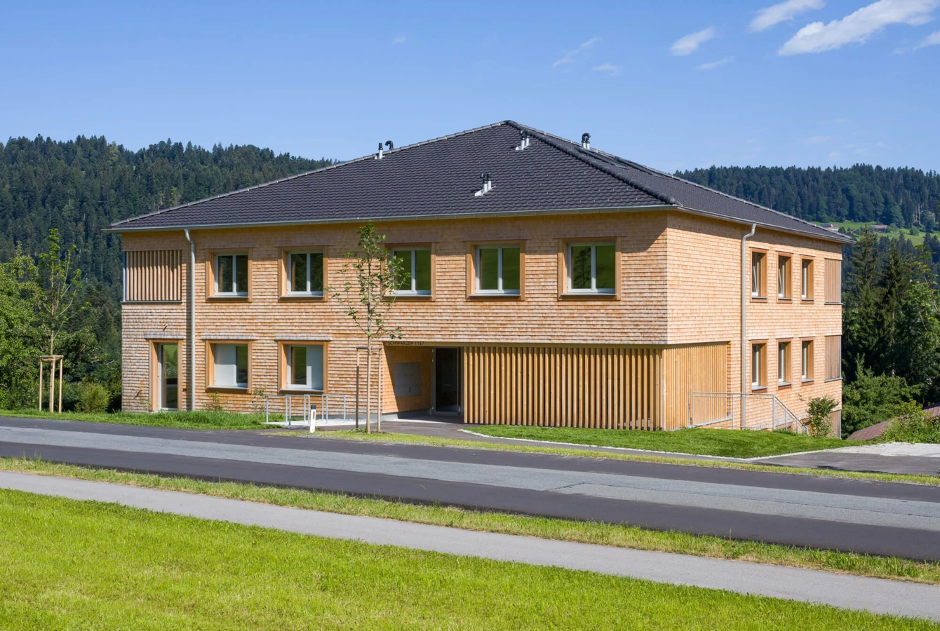 Modernes Wohnhaus mit Holzfassade und dunklem Dach, umgeben von grünen Wiesen und Wäldern unter blauem Himmel in Schwarzen, Alberschwende (Wohnanlage).