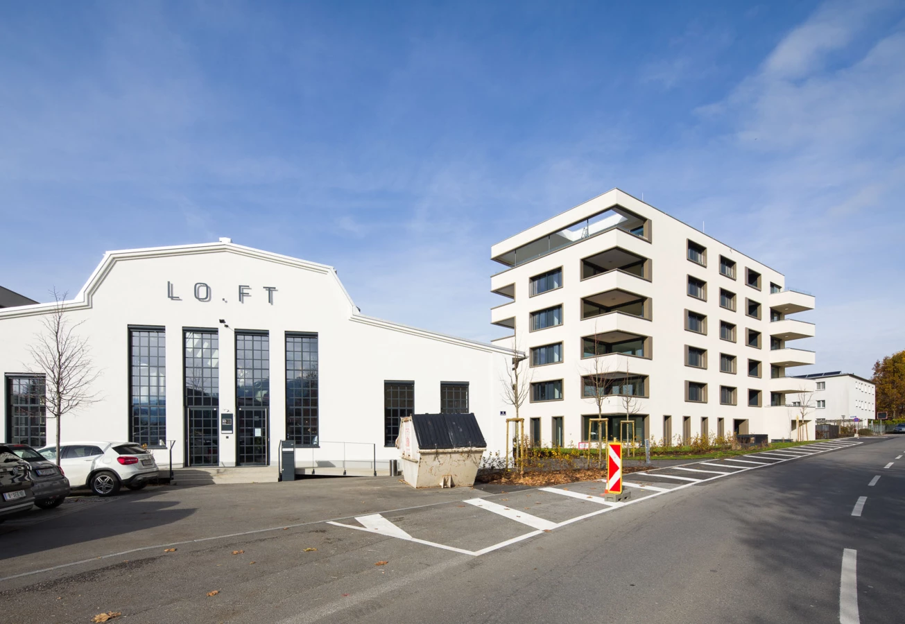 Zwei moderne Gebäude, eines mit "LO.FT" Schriftzug und großen Fenstern, sowie ein Wohnhaus mit Balkonen an einer Straße bei Tag (Bäumleareal).