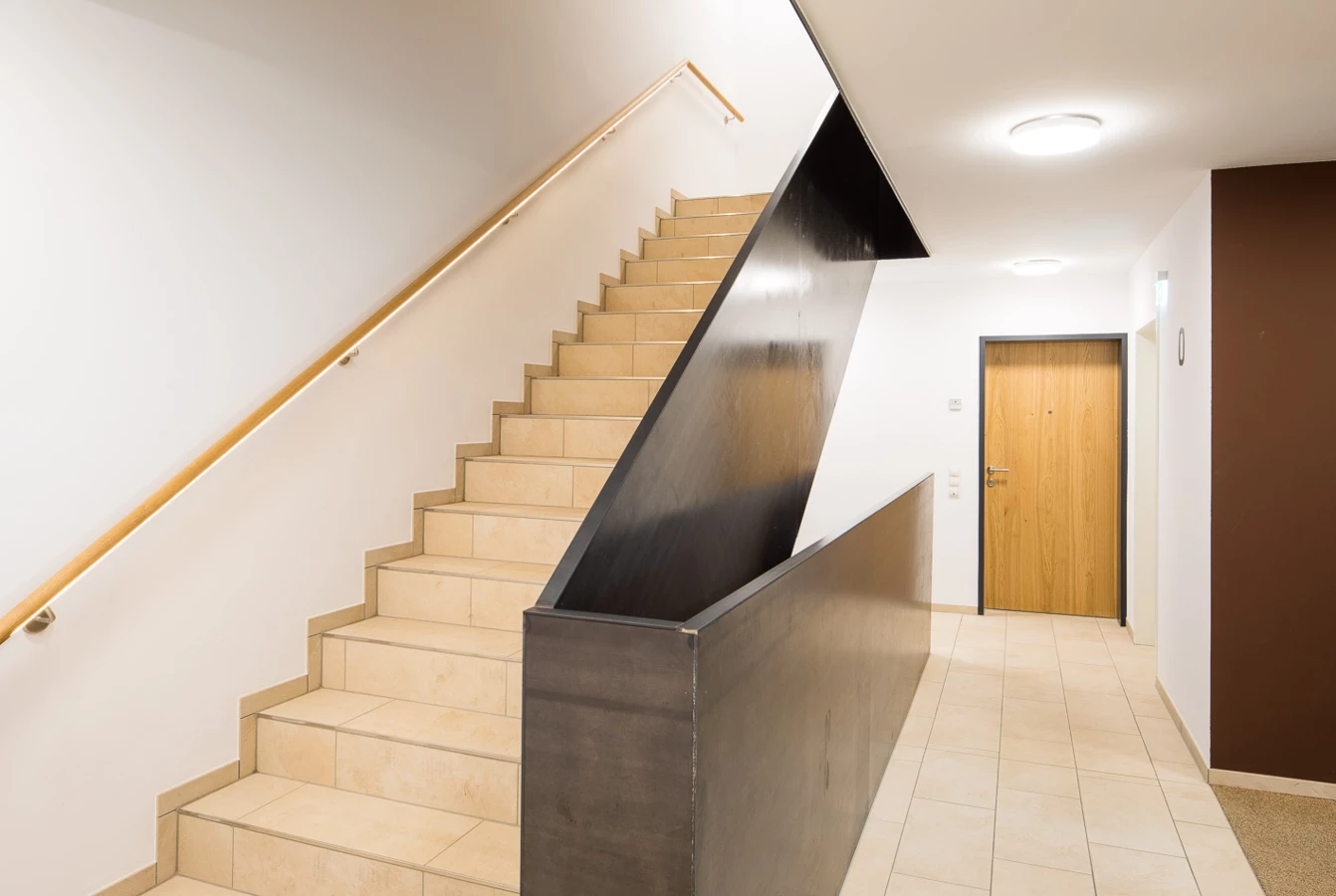 Ein modernes Treppenhaus mit hellen Fliesenstufen, einem Holzhandlauf und einem dunklen Metallgeländer. Eine Holztür ist am Ende des Flurs sichtbar (Baeumlequartier).
